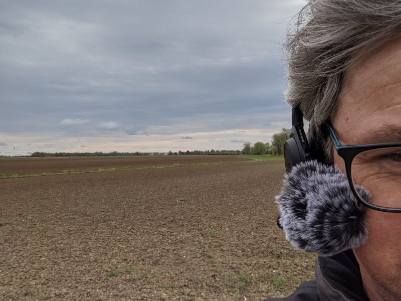 Das Bild zeigt ein Feld mit dem Kopf einer Person im Vordergrund. Das Gesicht der Person ist nur halb zu sehen. Die Person trägt ein Paar Kopfhörer mit einem pelzigen Mikrofon daran befestigt. Das Feld ist weitläufig und karg mit einigen Bäumen im Hintergrund. Der Himmel ist bewölkt und bedeckt. Das Bild scheint aus einem niedrigen Winkel aufgenommen worden zu sein.